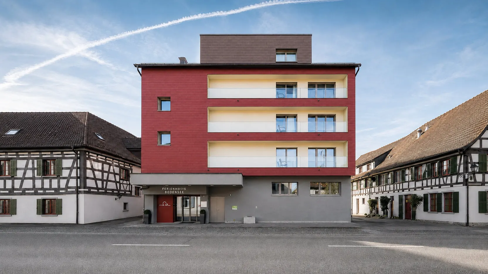 Gesamtansicht des Ferienhotels Bodensee in Berlingen mit roter Fassade, geschützten hellen Loggien und den historischen Fachwerkhäusern der Nachbarschaft.