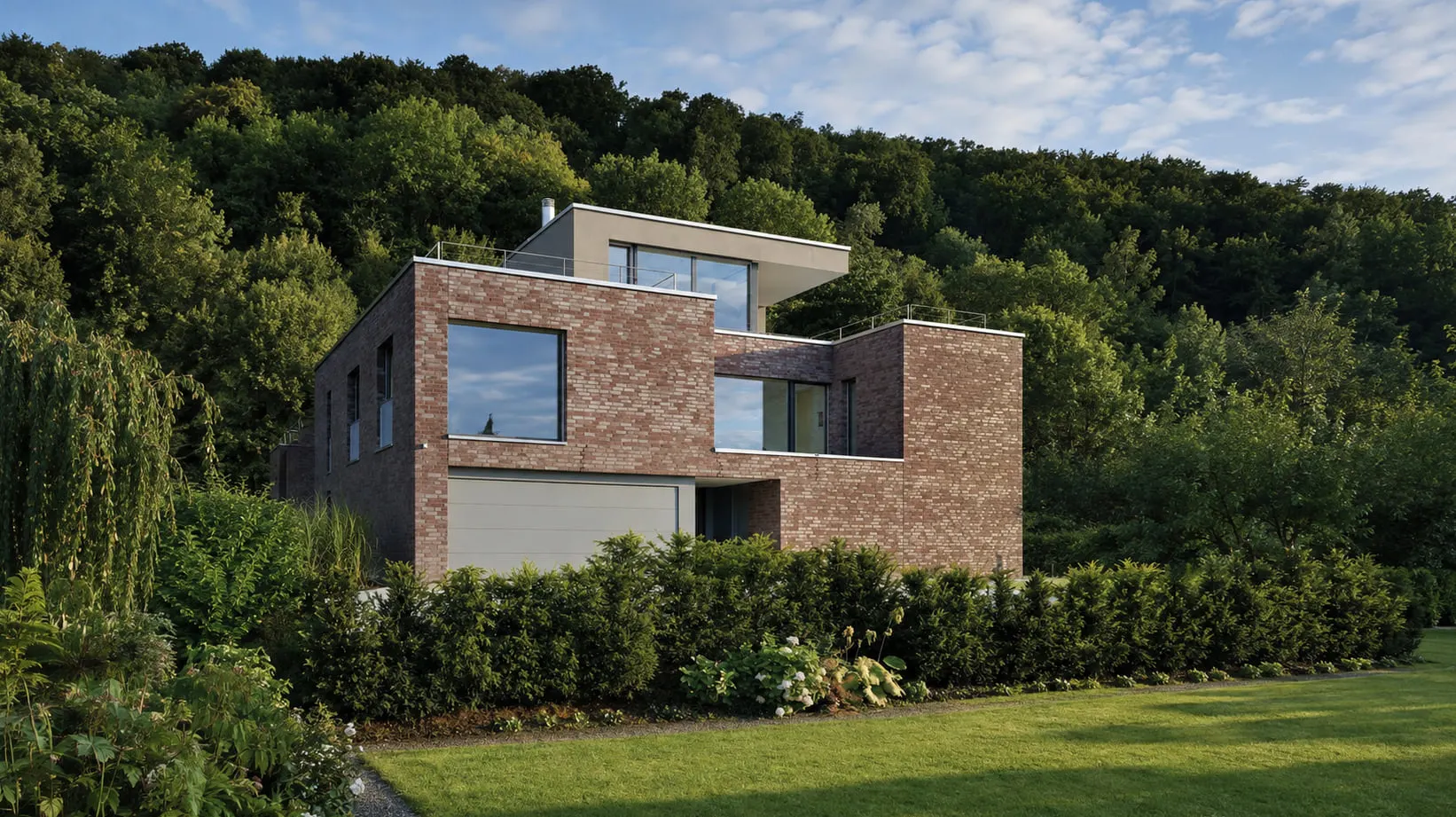 Gartenansicht des Hauses am Waldrand mit versetzten Ziegelbaukörpern vor dem Wald.
